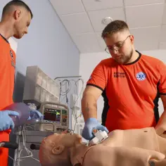 Dwóch ratowników medycznych ćwiczy resuscytację na fantomie, używając worka samorozprężalnego i defibrylatora.