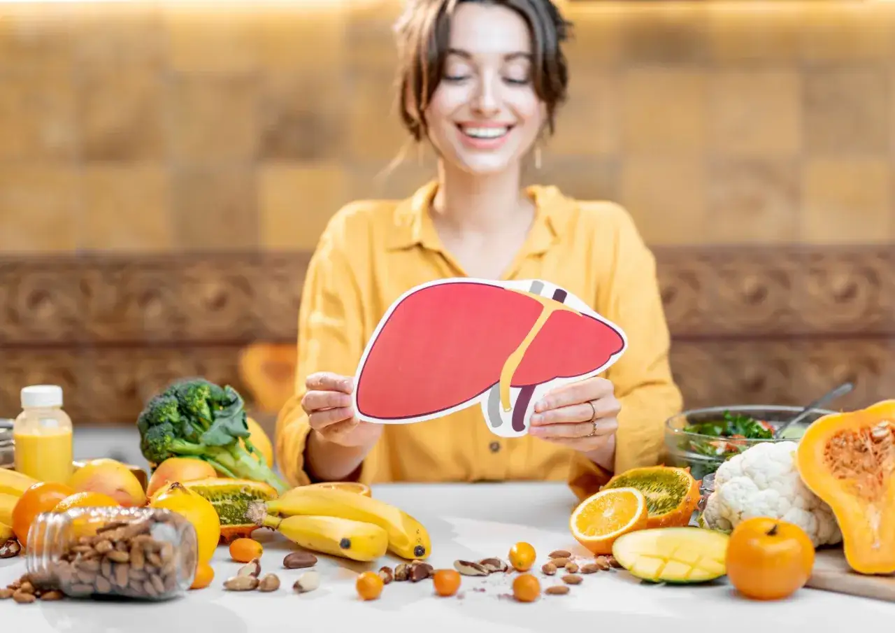 Uśmiechnięta kobieta trzyma model wątroby, otoczona owocami, warzywami i orzechami. Zdrowa dieta dla ggt.