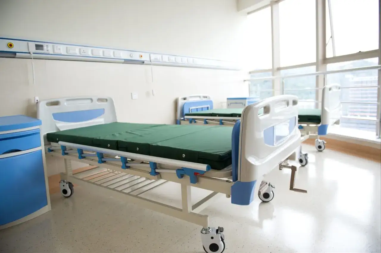 Szpitalne łóżka z zielonymi materacami w jasnym pokoju z oknami.